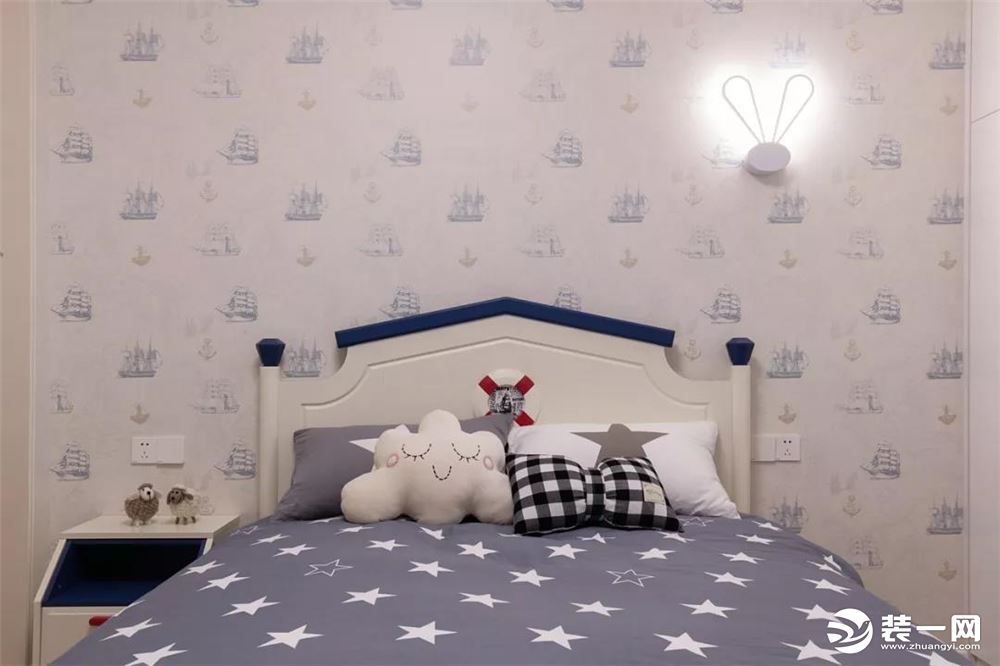 ▲床头墙是浅蓝色的船队航行图壁纸，搭配一盏兔耳床头壁灯，铺上一款满满繁星的被单，以云朵、蝴蝶结的抱枕