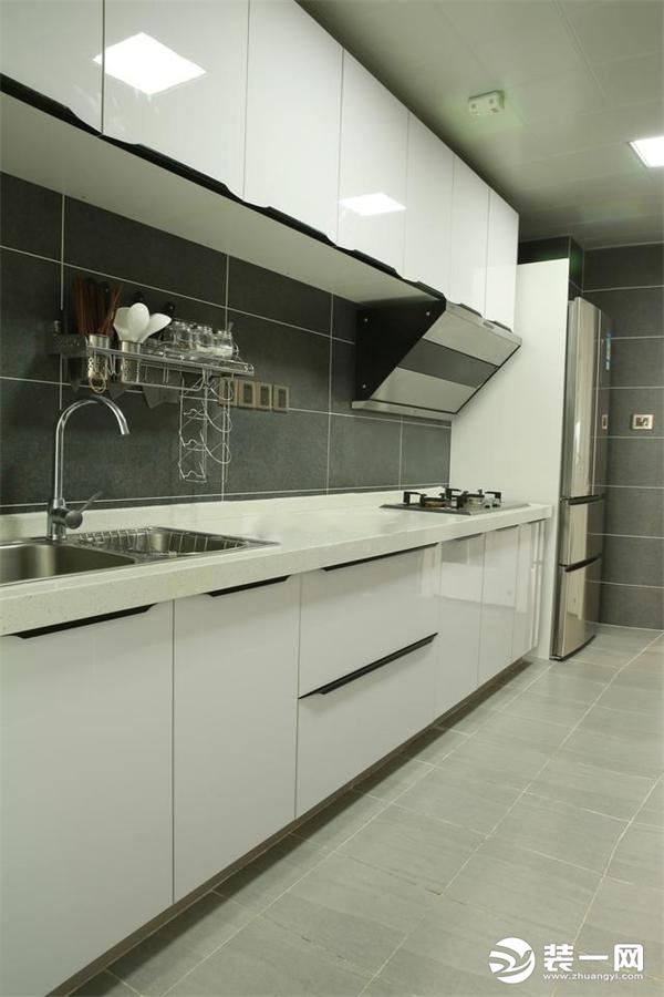 厨房是条形的空间，安装一字型白色整体橱柜，墙面黑色瓷砖，地面则是水泥灰的瓷砖，和整体色调优雅过渡