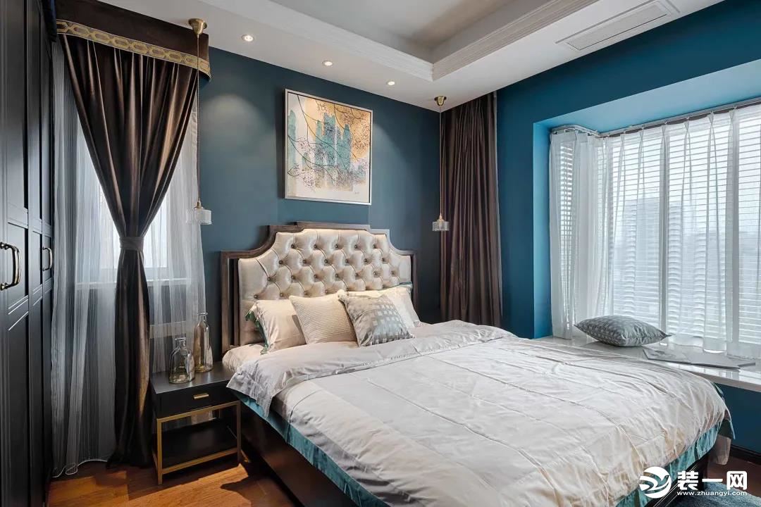 主卧在蓝色的墙面空间基础，搭配黑色的衣柜、床头柜，摆上布艺靠背的美式床，简素的床单与床头挂画，也是显
