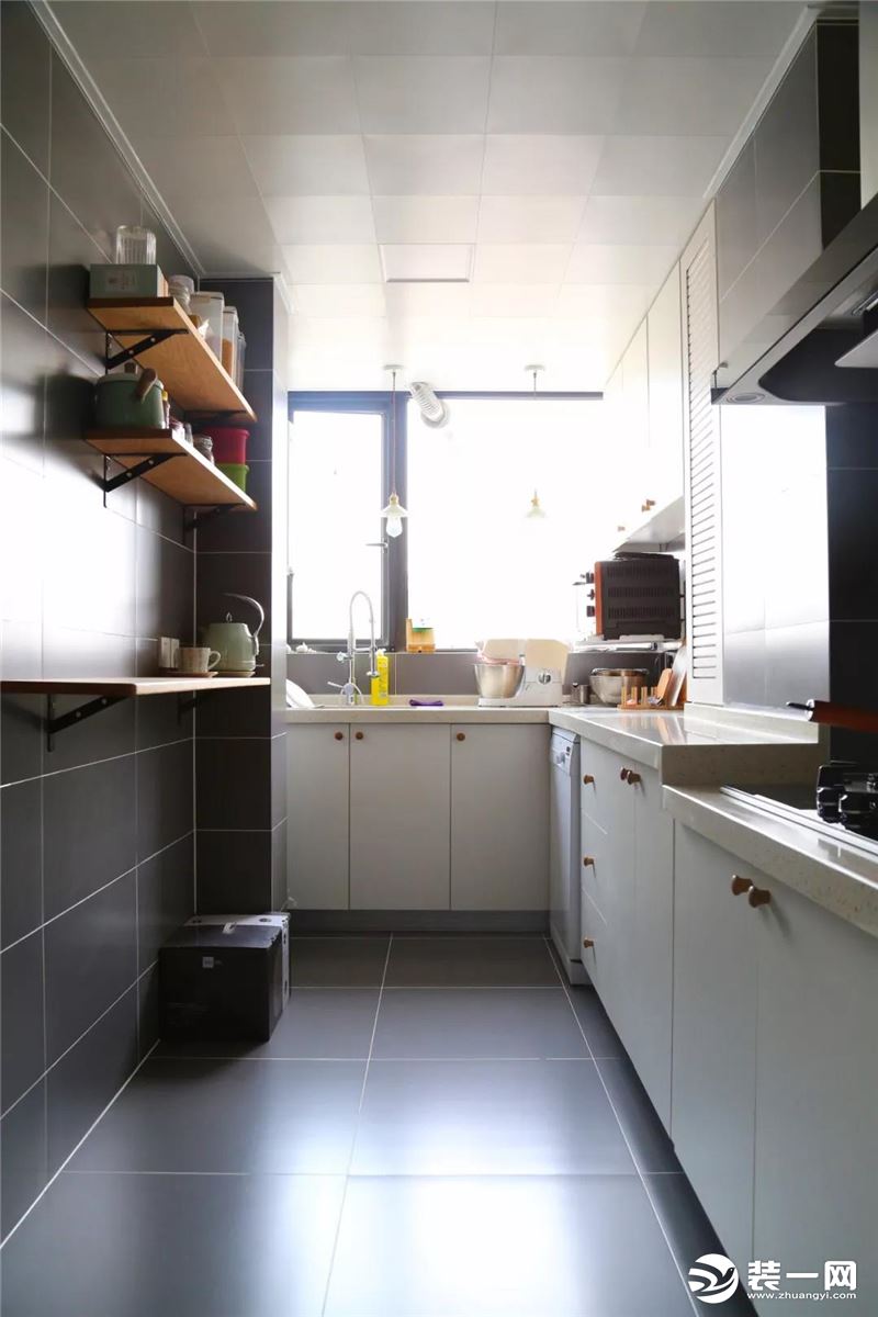 厨房内部是做了L形的橱柜布局方案，另一侧的墙面也做了隔板收纳设计。整个厨房以灰白色为主，深灰色的墙地