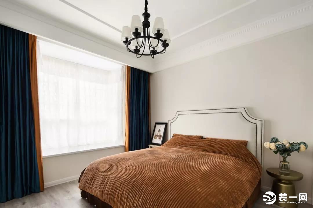 主卧干净的墙面基础氛围，造型优美的床头靠背搭配复古的驼色床品，与窗帘相呼应，而飘窗内测挂上橙色拼深蓝