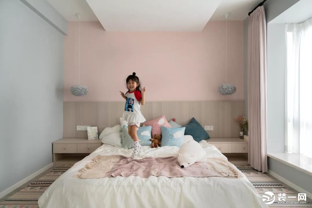 儿童房床头墙是粉色的，靠背的木质护墙板与床头柜连成一体式的，侧边墙面则是蓝灰色的，搭配上温馨可爱的床
