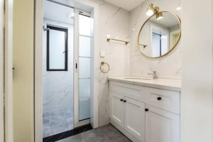 卫生间以白色大理石质感墙面，搭配简约灰色调的地面，整体白色的洗手盆柜，搭配一个圆形的金属框镜子