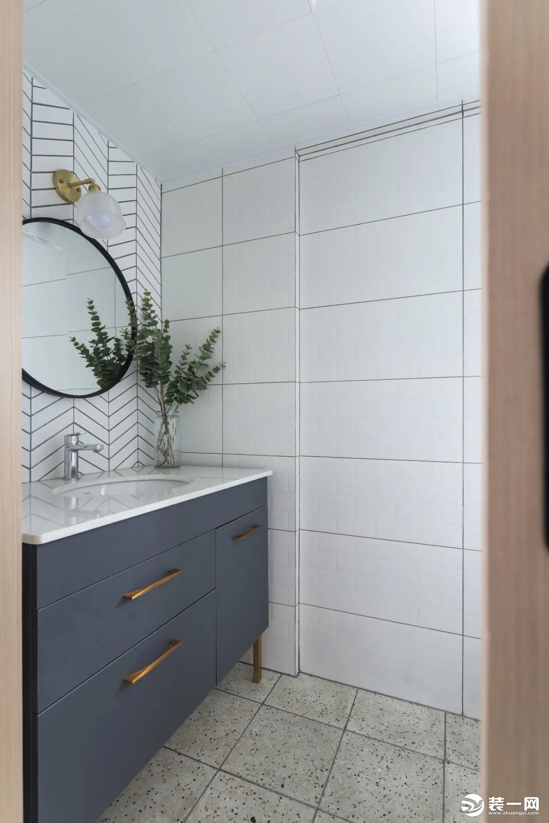  卫生间的水磨石地砖+白色墙砖，定制灰色洗手盆前方的墙面是鱼骨铺的墙面砖，挂一面圆形的镜子，让卫浴空