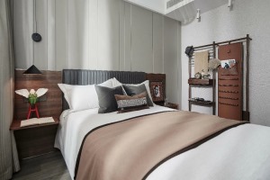 次卧的床头墙是灰色的硬包背景墙，床头靠背区域是木饰面材质，布置灰色皮艺的床铺，结合白色+咖色的床单，