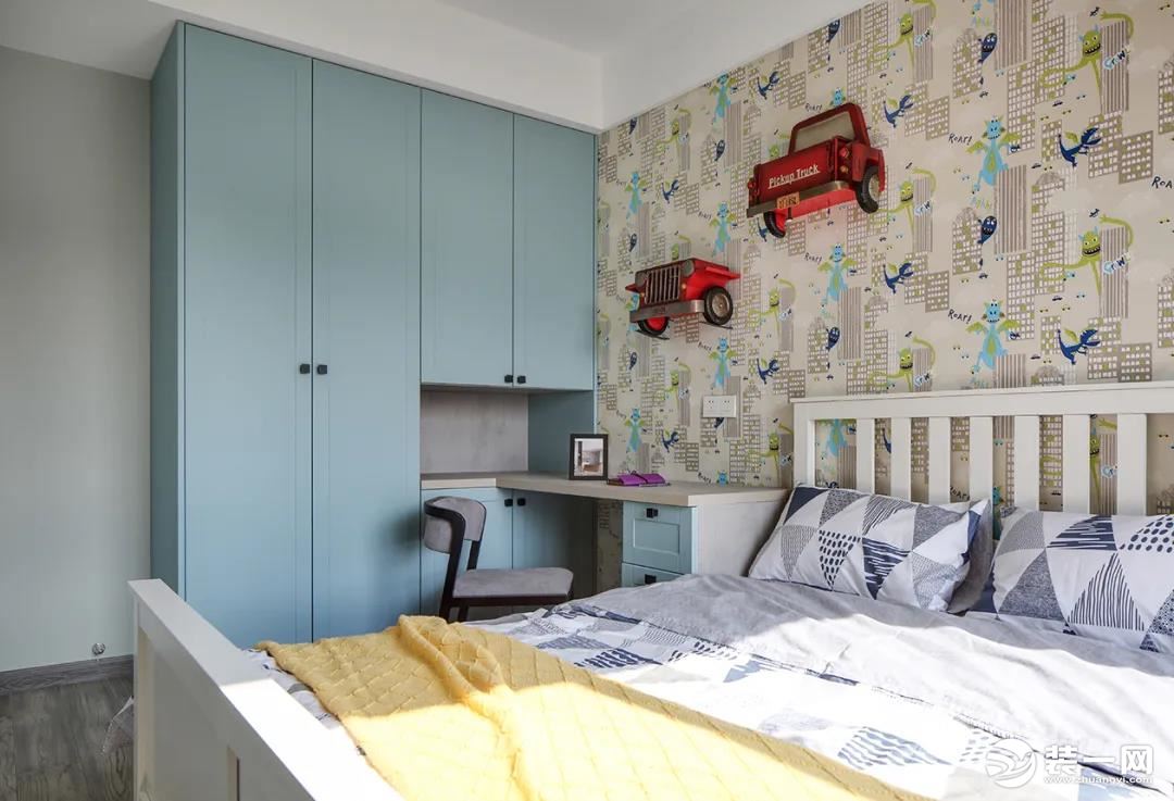 儿童房的床头墙是贴着卡通图案的墙布，上面挂着两个立体汽车小饰品，搭配淡蓝色的衣柜与抽屉柜、白色木栏靠