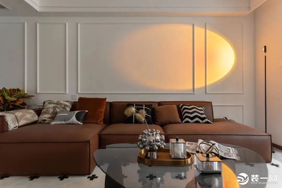布置棕色的皮质主体沙发，背景墙以简约的石膏线条勾勒，增强空间层次感，一盏落日灯映射于墙面，家居氛围瞬