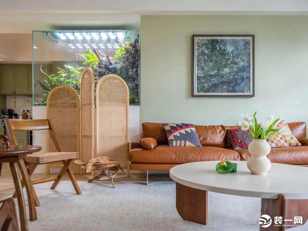 沙发背景墙选择浅绿色，以艺术画像点缀装饰，搭配棕色复古皮艺沙发，色彩层次丰富，一侧藤编元素的屏风为空