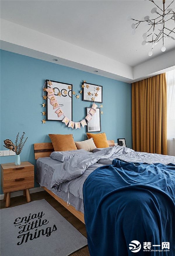 木质地板舒适温暖，灰色床品与地毯，深蓝色的毛毯舒适惬意，淡蓝色的床头背景和几幅简单的挂画，枝蔓状的灯