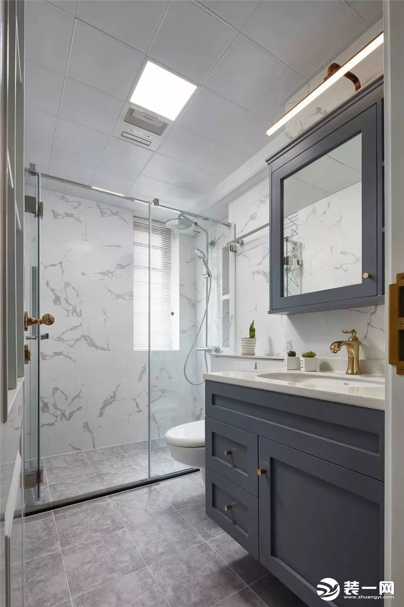 卫生间内用灰色地砖搭配白色大理石墙砖，再加上蓝灰色的浴室柜、浴室镜和精致的金色龙头、柜门拉手，让卫生